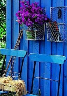 two blue chairs and some purple flowers on the side of a building in front of a blue wall