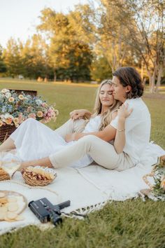 two women are sitting on a blanket in the grass with food and drinks around them