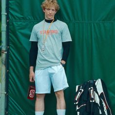 a young man standing next to a green wall holding a tennis racquet and bag