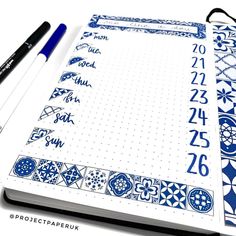 an open notebook with blue and white designs on it, next to two black pens