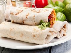 two burritos on a white plate next to some tomatoes and limes in a jar