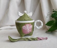 a ceramic cup with angel wings on it next to a pink ribbon and potted plant