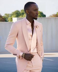 a man in a pink suit and gold bracelets is standing on the street with his hands in his pockets