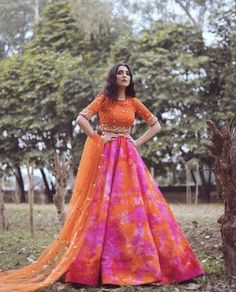 a woman in an orange and pink dress standing next to a tree with her hands on her hips