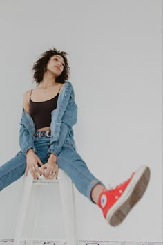 a woman sitting on top of a white stool wearing red converses and denim jacket