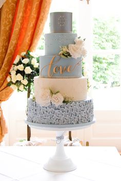 a three tiered cake with flowers on top is displayed in front of a window