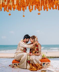 a bride and groom sitting on a bench under an orange flowered canopy in front of the ocean