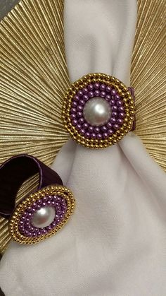 two pieces of jewelry sitting on top of a white cloth covered table next to a gold fan