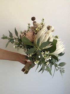 a person holding a bouquet of white flowers and greenery in their hand, against a white wall