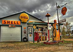 an old fashioned gas station sits empty on the side of a dirt road in front of a cloudy sky