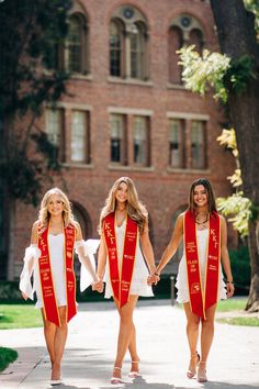 three women in graduation gowns holding hands and walking down the street with their robes on