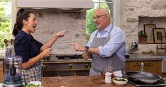 an older man and woman in the kitchen talking to each other while pointing at something