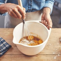 a person whisking eggs in a white bowl on a wooden table with utensils