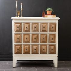 a white dresser with wooden drawers and candles on top, against a black wall behind it