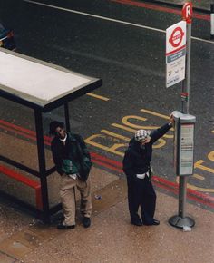 two people standing next to a bus stop on the side of the road with their hands in the air