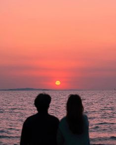 two people watching the sun set over the ocean in front of an orange and pink sky