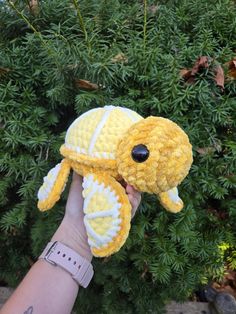 a hand holding a yellow stuffed animal in front of a bush with green leaves on it