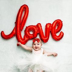 a baby in a tutu laying on the floor next to balloons that spell out love