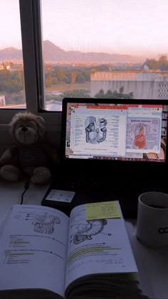 an open book sitting on top of a desk next to a cup and laptop computer