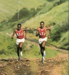 two men running down a dirt road with grass and hills in the backgroud