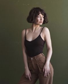 a woman standing in front of a green wall wearing brown pants and a black tank top