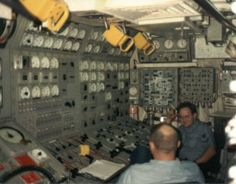 two men sitting in the control room of a ship looking at something on the wall