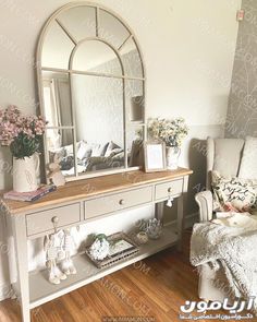 a mirror sitting on top of a dresser next to a white chair and flower vase