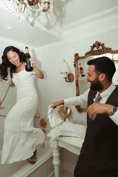 a woman in a white dress is holding a wine bottle and standing next to a man