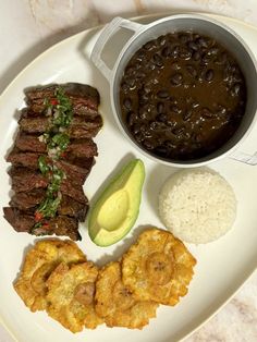 a white plate topped with meat, rice and beans next to a bowl of beans