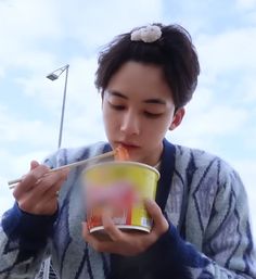 a young man eating noodles from a cup with chopsticks in his hand while holding a drink