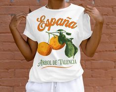 Show off your love for citrus with this eye-catching Spanish orange tee featuring retro orchard imagery. A vintage illustration of Valencia oranges and branches is framed by the Spanish words 'España' and 'Árbol de Valencia'. Celebrating the sweet, juicy Valencia oranges from the Mediterranean climate of Spain, this graphic tee is perfect for citrus lovers and world travelers alike. The bright, sunny design evokes lazy afternoons under Orange tree groves in Valencia. Crafted from soft cotton in Orange Printed Graphic Tee, Retro Orange T-shirt For Spring, Vintage Fruit Print Crew Neck Tops, Vintage Crew Neck Tops With Fruit Print, Valencia Orange, Orange Tees, Spanish Words, Orange T Shirts, Orange Tree