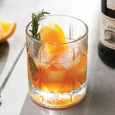 an orange drink with ice and rosemary garnish in a glass next to a bottle