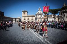 a group of men in roman garb marching down a street with people standing around