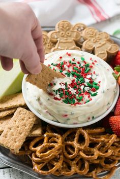 a person dipping crackers into a bowl of dip surrounded by pretzels and strawberries