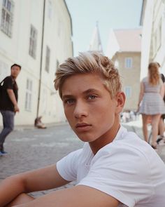 a young man with blonde hair sitting on the ground in front of some buildings and people