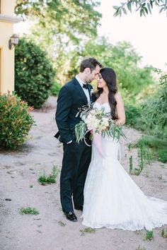 a bride and groom standing in front of a house with greenery on the ground