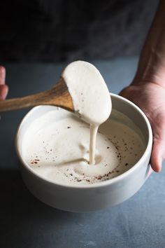 a person holding a wooden spoon over a bowl filled with yogurt and spices