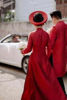 two people in red dresses and hats walking down the street with a white car behind them