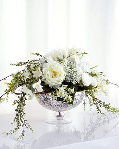 a white flower arrangement in a silver bowl on a glass table with curtains behind it