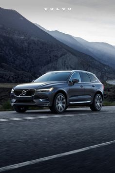 the volvo suv is driving down the road in front of some mountains and water, while another car is parked on the side of the road