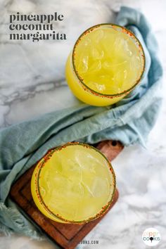 two glasses filled with pineapple margarita sitting on top of a wooden cutting board next to a blue towel