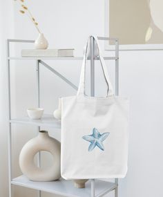 This aesthetic coastal tote bag is ready for a trip to the beach, or to carry your book to your favorite coffee shop. This is versatile as a book bag, beach bag, or errand tote. This watercolor painting of a starfish is printed on a sustainable cotton canvas tote bag, making for a minimalist addition to your summer look! Details: - One size: 15" x 16"  - 20" handle length  - 100% cotton canvas tote - Lightweight and compact - Print is on one side, the other side is blank  - The reinforced handle Eco-friendly Canvas Bag For Beach, Eco-friendly Canvas Bag For The Beach, White Cotton Canvas Bag For Beach Season, White Cotton Canvas Bag For Vacation, Eco-friendly Cotton Canvas Beach Bag, Rectangular Canvas Bag With Eco-friendly Ink For Summer, Eco-friendly White Canvas Beach Bag, Eco-friendly Ink Canvas Bag For Everyday Summer Use, Beachy Cotton Tote Bag