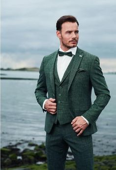 a man wearing a green suit and bow tie standing next to the ocean with his hands in his pockets