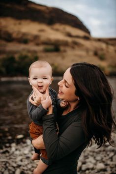 a woman holding a baby up to her face while standing next to a body of water