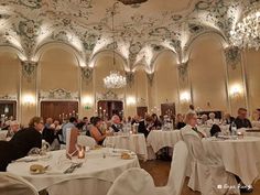 many people are sitting at tables with white linens and chandeliers on the ceiling