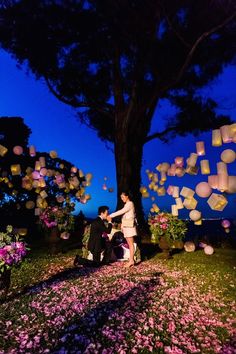 two people standing under a tree with paper lanterns flying in the air above them at night