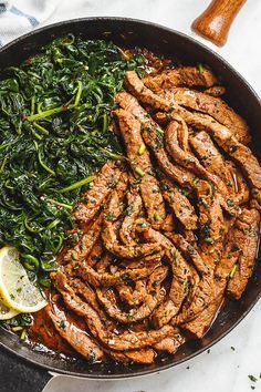 a skillet filled with meat, greens and lemon wedges next to a wooden spatula