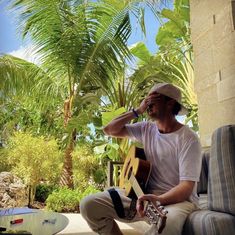 a man sitting on a couch with a guitar in his hand and palm trees behind him