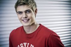 a young man wearing a red tshirt smiles at the camera while standing in front of a metal wall