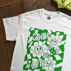 "Grow With The Flow Graphic T-Shirt - Oversized Unisex White Tee - 100% Cotton, Eco-Friendly - Get Gardening In Style White cotton tee illustrated with a green graphic print of smiley flowers, wavey checkerboard pattern and a cheery quote of \"Grow With The Flow\".  🌻 unisex t-shirt 🧵 100% cotton 📏 relaxed fit ⚖ midweight fabric 203 gsm 🌍 planet friendly 💛 sustainably sourced materials  💧 printed using vegan friendly, water-based inks  🌟 made to order, printed just for you! (a more sustainable method that avoids unnecessary textile waste) 🐳 posted in a plastic free biodegradable mailing bag ♻ packaging is 100% recyclable after use ©2024 Kio Cards Ltd. All images, artwork and content. All rights reserved. say hi/tag us on Instagram @sixsunflowersdesign" Flow Illustration, Stud Aesthetic, Festival Tshirt, Green Graphic Tee, Grow With The Flow, Flow Graphic, Checkerboard Pattern, Garden Kitchen, Typography Tshirt